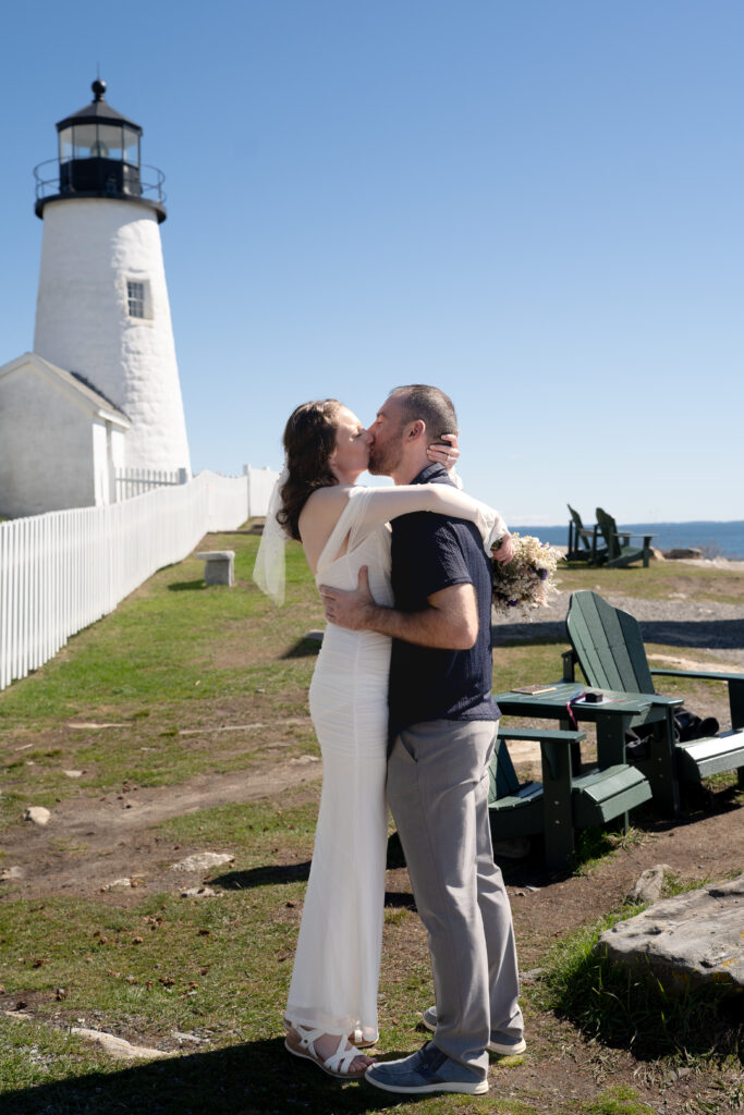 Couple holding hands during elopement vows beside Pemaquid Point Lighthouse surrounded by Maine coastline in bloom