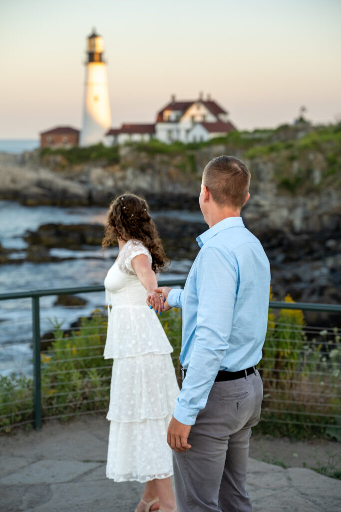 Bride and groom standing on rocky cliffs beside Portland Head Light during a summer elopement with bright ocean views