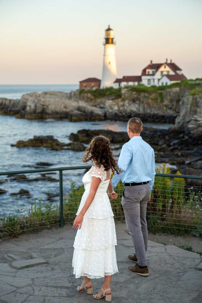 Romantic couple portrait at Portland Head Light during golden summer sunset with waves crashing below the cliffs